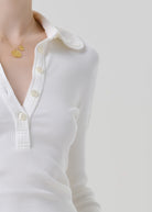 Close-up of a person wearing a white ribbed long-sleeve polo-style top with a collar and white buttons, paired with a delicate gold necklace, against a light gray background.