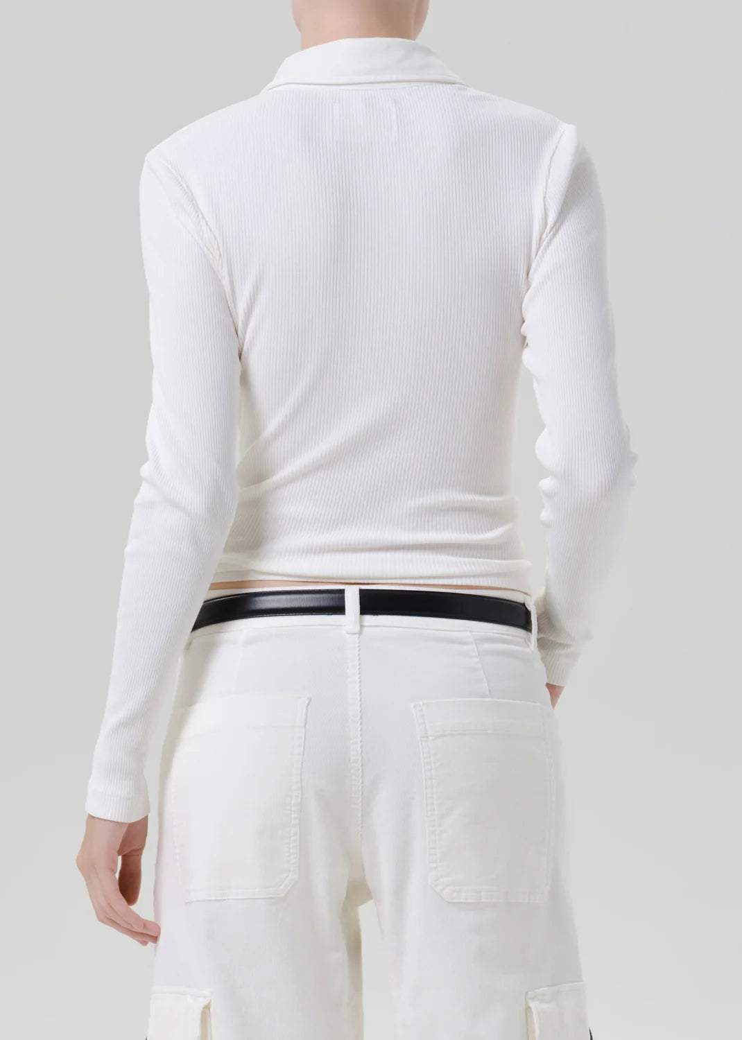 A model shown from the back wearing a white ribbed long-sleeve polo shirt tucked into white pants with a thin black belt.