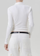 A model shown from the back wearing a white ribbed long-sleeve polo shirt tucked into white pants with a thin black belt.