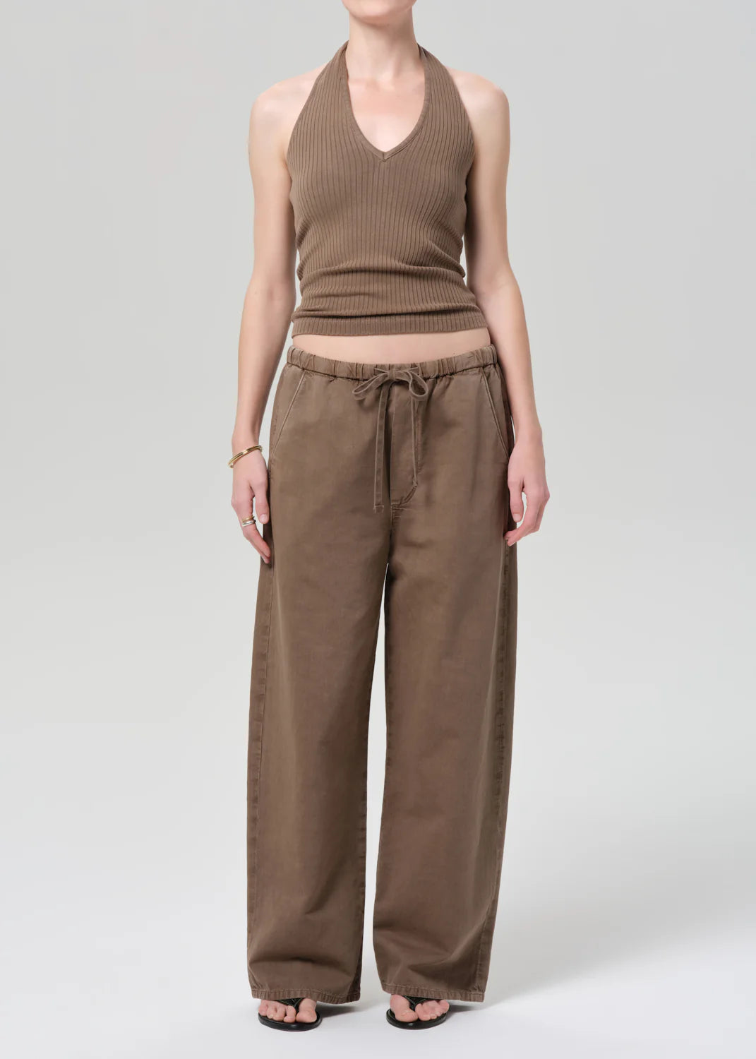 Person wearing a brown ribbed tank top and brown pants on a gray background with flip flops.