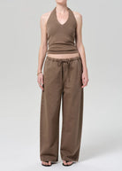 Person wearing a brown ribbed tank top and brown pants on a gray background with flip flops.