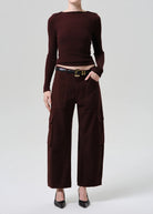 A model wearing a dark brown long-sleeve knit top and matching dark brown wide-leg cargo pants, styled with a black leather belt and black pointed-toe heels against a light gray studio background.