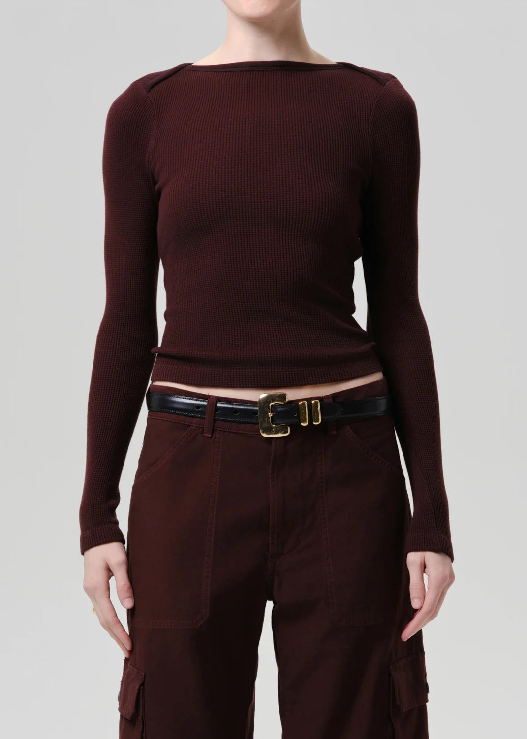 A model wearing a dark burgundy ribbed long-sleeve top with shoulder details, paired with matching dark brown cargo pants and a black belt with a gold buckle, against a solid light gray background.