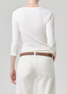 Person wearing a white long-sleeve top and white pants with a brown belt on a plain background