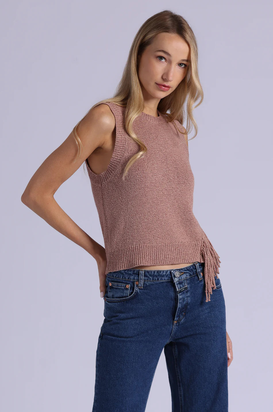 Woman wearing a sleeveless brown knit top with fringe and blue jeans on a light gray background