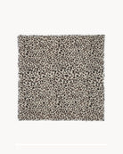 A square textile swatch with a dense leopard print pattern in black and dark brown on a warm beige background, finished with short fringe on all sides.