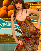 Woman in a floral dress standing in front of a scenic background with buildings and water.
