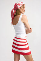 Woman wearing a white tank top and red and white striped skirt with a headscarf on a plain background