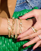 A close-up of a person's hands and wrist wearing multiple gold-toned chain bracelets and rings, with dark red nail polish, a brown leather sleeve, blue denim, and a bright green quilted bag in the background.