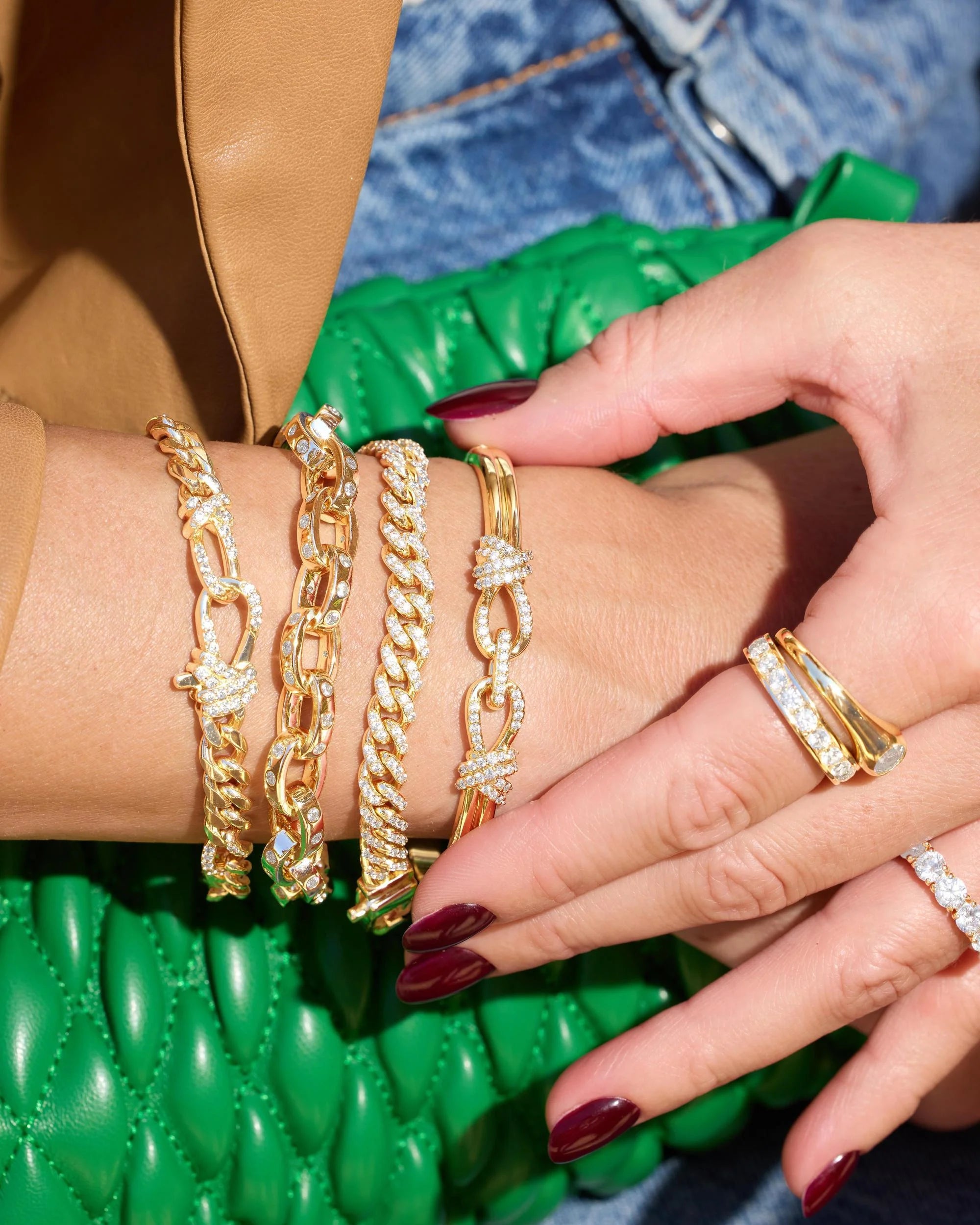 A close-up of a person's hands and wrist wearing multiple gold-toned chain bracelets and rings, with dark red nail polish, a brown leather sleeve, blue denim, and a bright green quilted bag in the background.