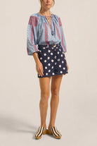 Woman wearing a blue embroidered blouse and navy polka dot skirt on a beige background