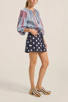 Woman wearing a blue embroidered blouse and navy polka dot skirt on a beige background