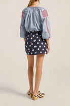 Woman wearing a blue blouse with red embroidery and a navy floral skirt on a beige background