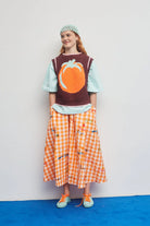 Person wearing a brown sweater with a pumpkin design, light blue shirt, orange checkered skirt, and green sneakers on a white background.