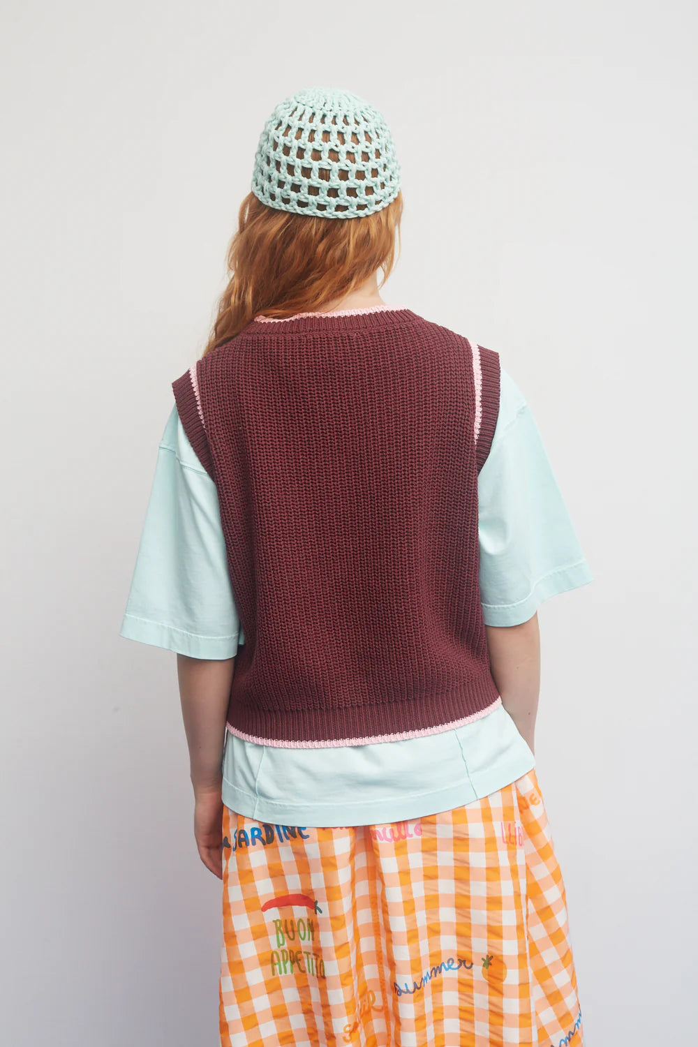 Person wearing a maroon knitted vest over a light blue shirt with orange checkered pants on a plain background