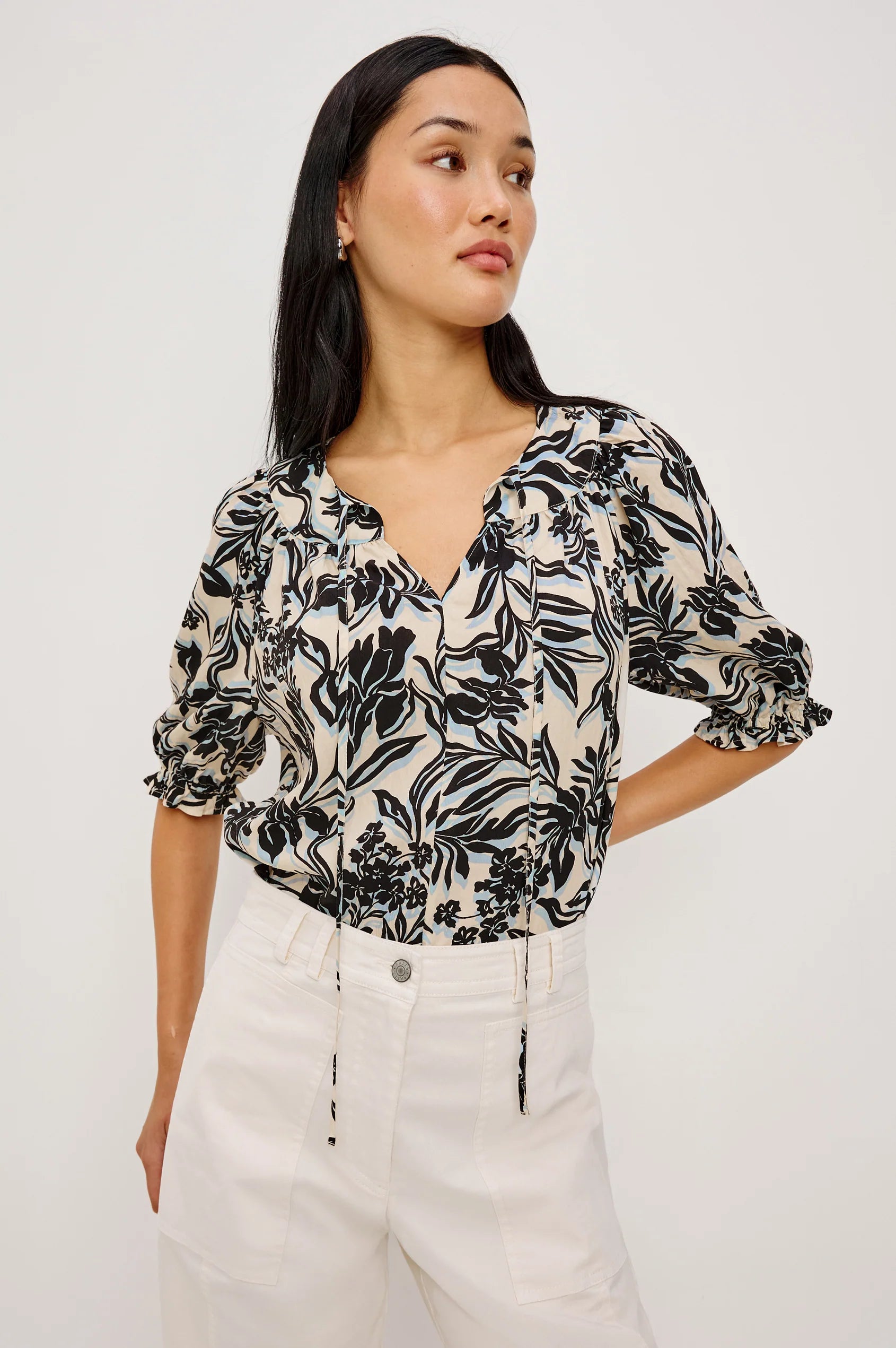 Woman wearing a black floral blouse and white pants against a plain background