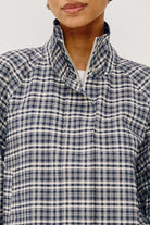 A close up of a woman modeling a navy and white checkered long-sleeve cropped jacket with a high stand-up collar, zipped all the way up, against a plain light gray background.