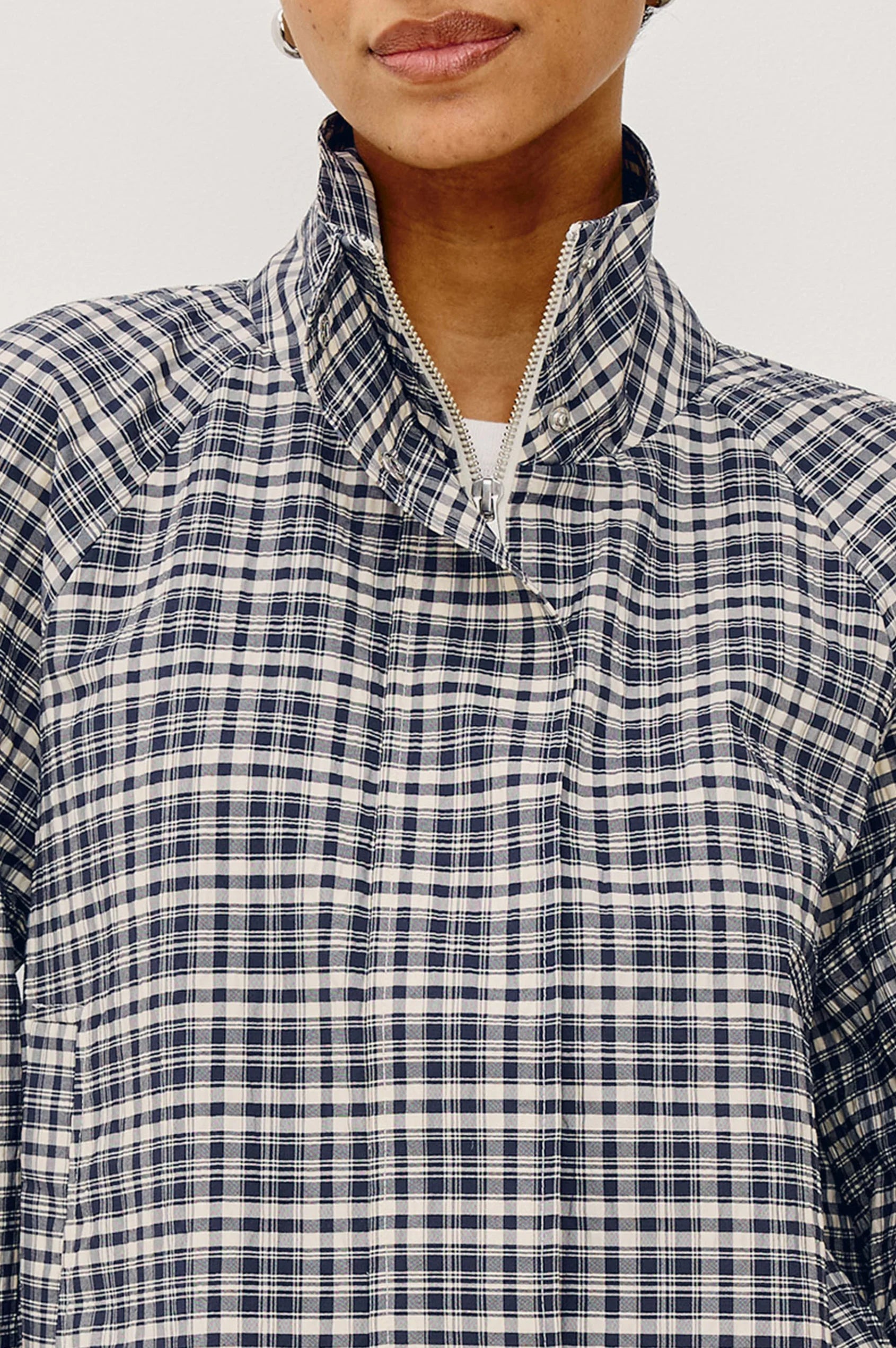 A close up of a woman modeling a navy and white checkered long-sleeve cropped jacket with a high stand-up collar, zipped all the way up, against a plain light gray background.