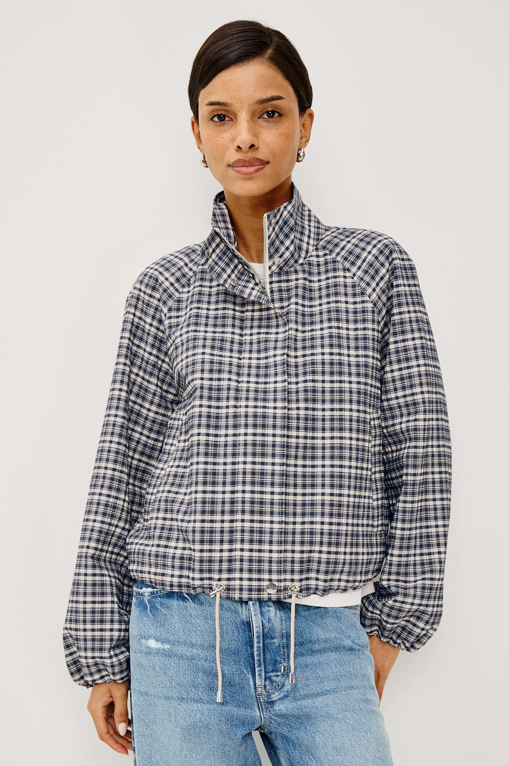 A woman modeling a navy and white checkered long-sleeve cropped jacket with a high stand-up collar, paired with light blue jeans, against a plain light gray background.