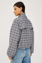A woman shown from the back wearing a cropped, puffer-style plaid jacket in a gray, white, and black check pattern, paired with blue jeans, against a plain light gray studio background.