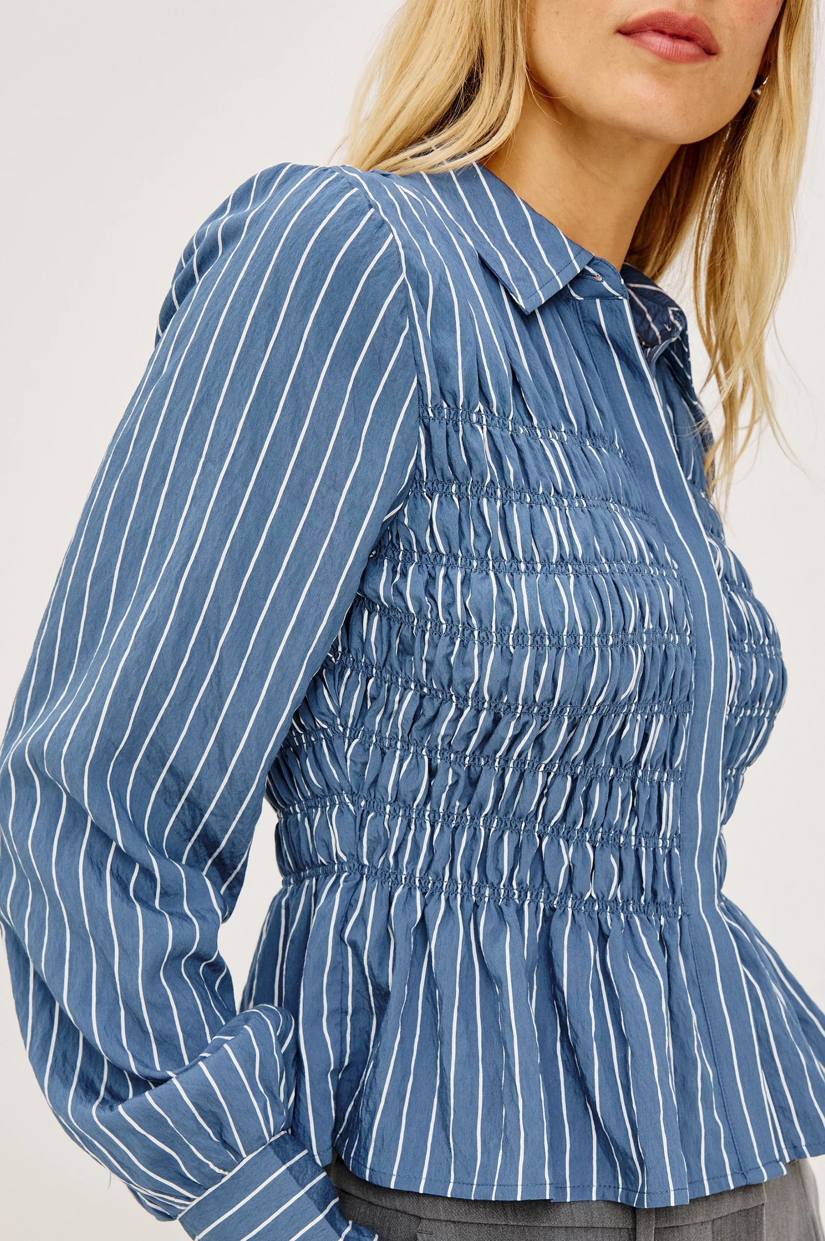 A close-up of a woman wearing a blue long-sleeve blouse with white vertical pinstripes and a smocked bodice, cropped from the mouth down against a plain light background.