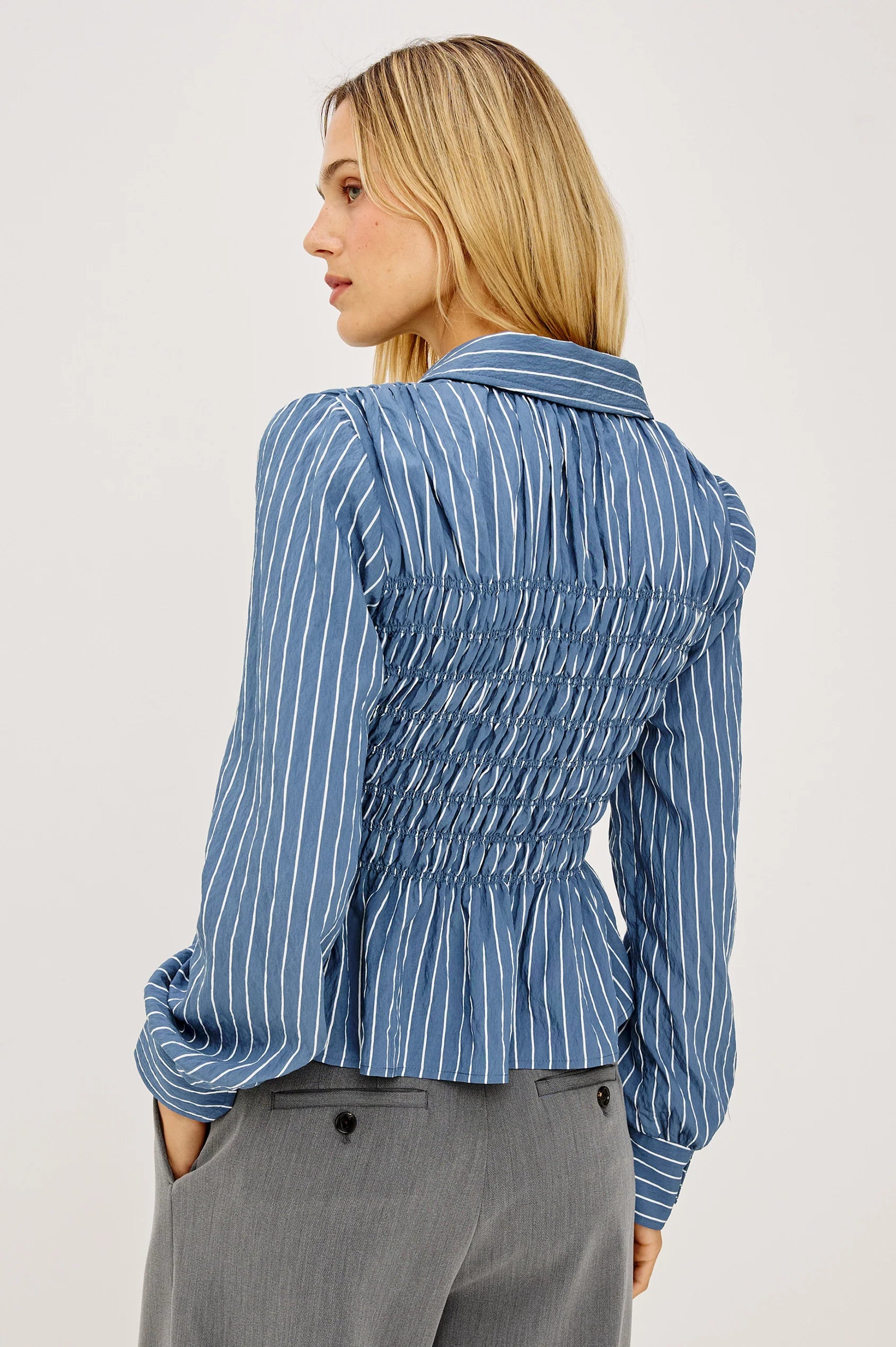 A woman shown from the back wearing a blue and white vertically striped long-sleeve blouse with a smocked waist and peplum hem, paired with gray trousers, against a plain light gray studio background.