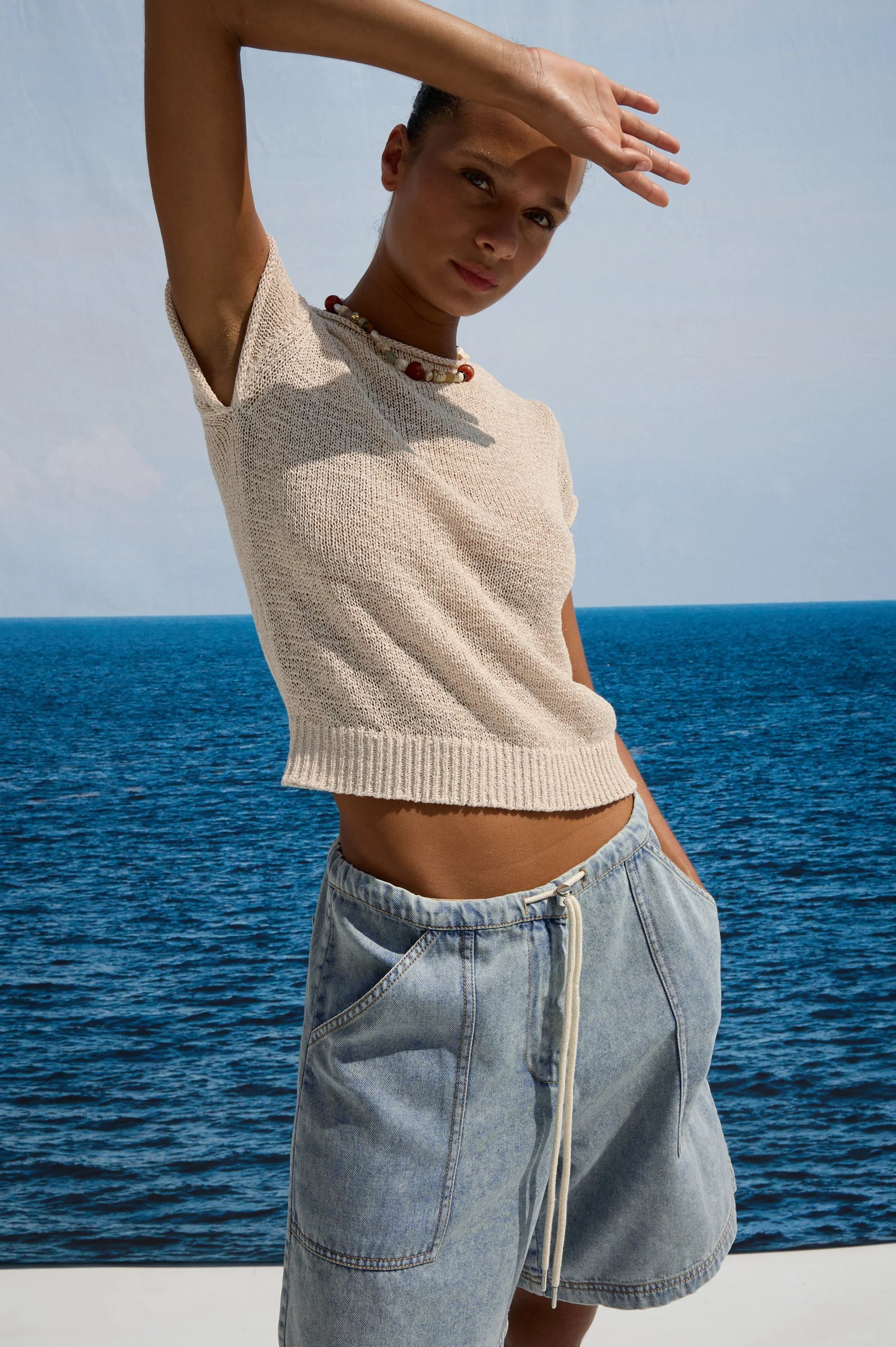 Woman in a beige top and light blue shorts standing by the ocean.