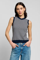 A woman modeling a sleeveless knit top with a navy and white geometric pattern, paired with blue jeans, against a solid light gray background.