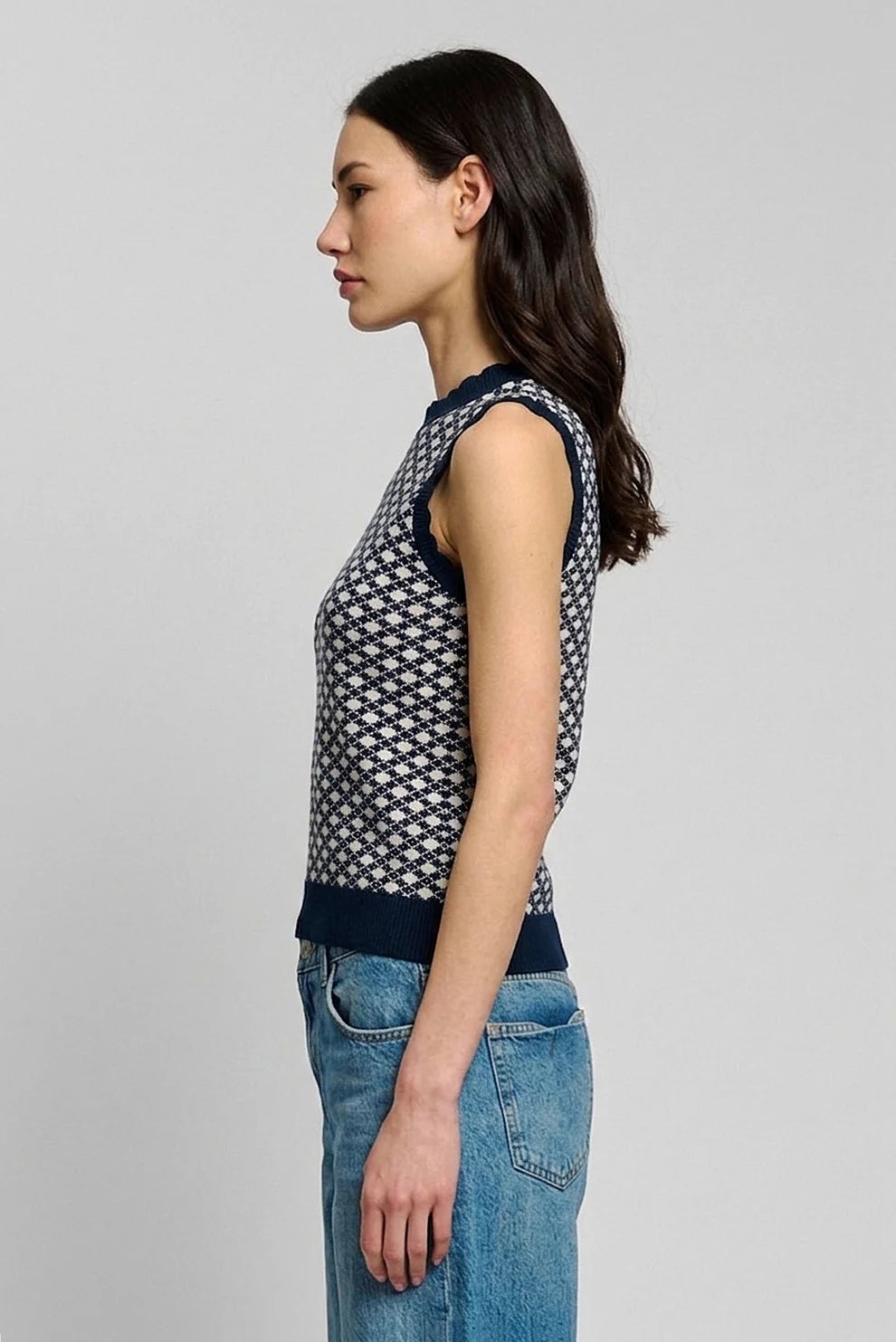 A woman shown in side profile wearing a sleeveless knit top with a navy and white geometric pattern, paired with blue jeans, against a plain light gray studio background.