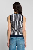 A woman shown in back profile wearing a sleeveless knit top with a navy and white geometric pattern, paired with blue jeans, against a plain light gray studio background.