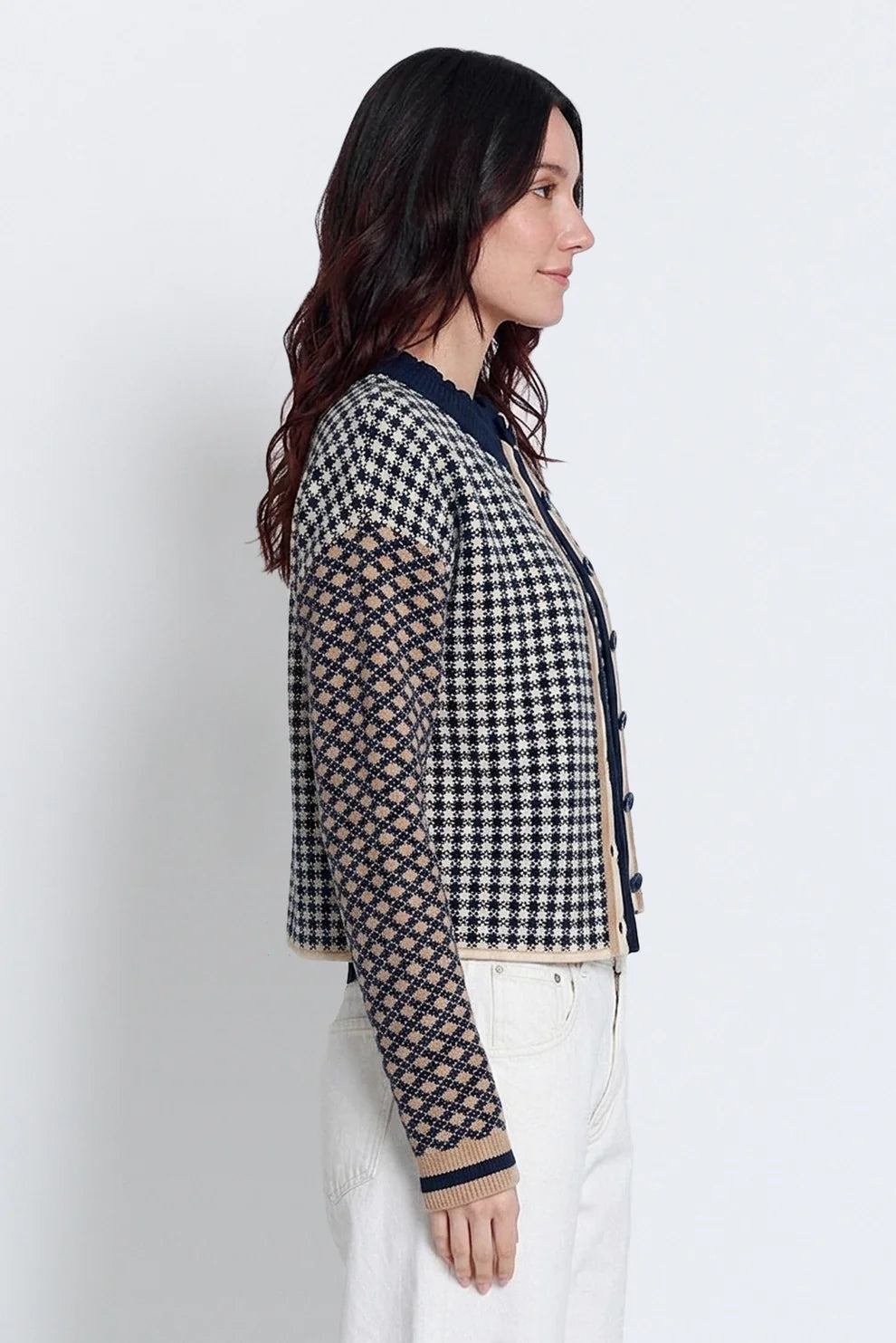 A woman shown in profile wearing a cropped houndstooth cardigan with patterned sleeves and white pants, photographed against a plain light gray studio background.