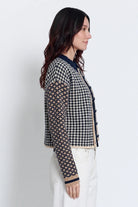 A woman shown in profile wearing a cropped houndstooth cardigan with patterned sleeves and white pants, photographed against a plain light gray studio background.
