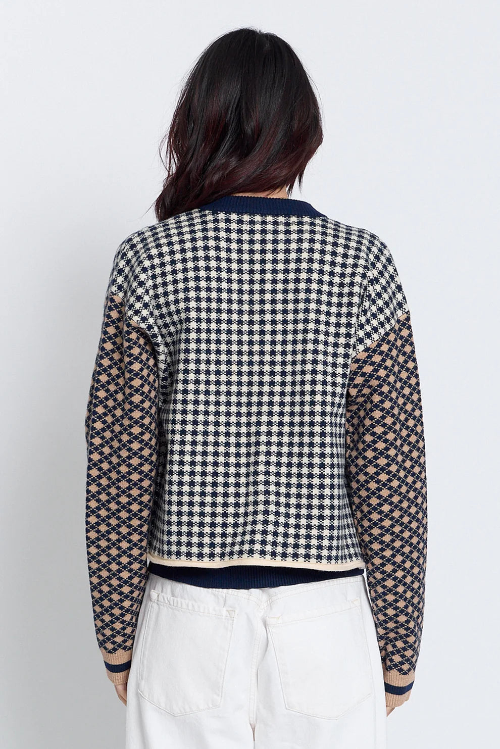 A woman viewed from the back wearing a cropped knit cardigan with a black and white houndstooth pattern on the body and sheer patterned sleeves, paired with white pants against a light gray studio background.