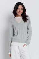 A woman wearing a light gray v-neck knit sweater over a white mesh top, paired with white pants, photographed against a light gray studio background.