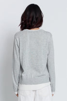 A person shown from the back wearing a light heather gray long-sleeve top with a white undershirt and white pants, against a plain light gray studio background.