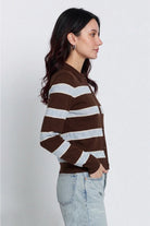 A woman in side profile wearing a brown and light gray striped knit cardigan sweater with light-wash jeans, photographed against a plain light gray studio background.