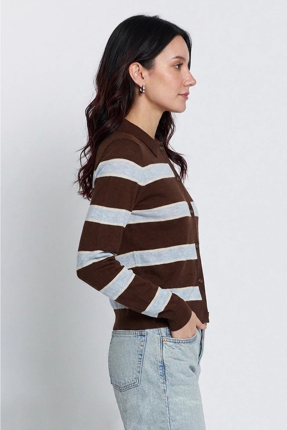 A woman in side profile wearing a brown and light gray striped knit cardigan sweater with light-wash jeans, photographed against a plain light gray studio background.