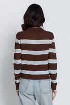 A woman viewed from the back wearing a brown and light gray striped long-sleeve knit top with a mock neck, paired with light-wash jeans, against a plain light gray studio background.