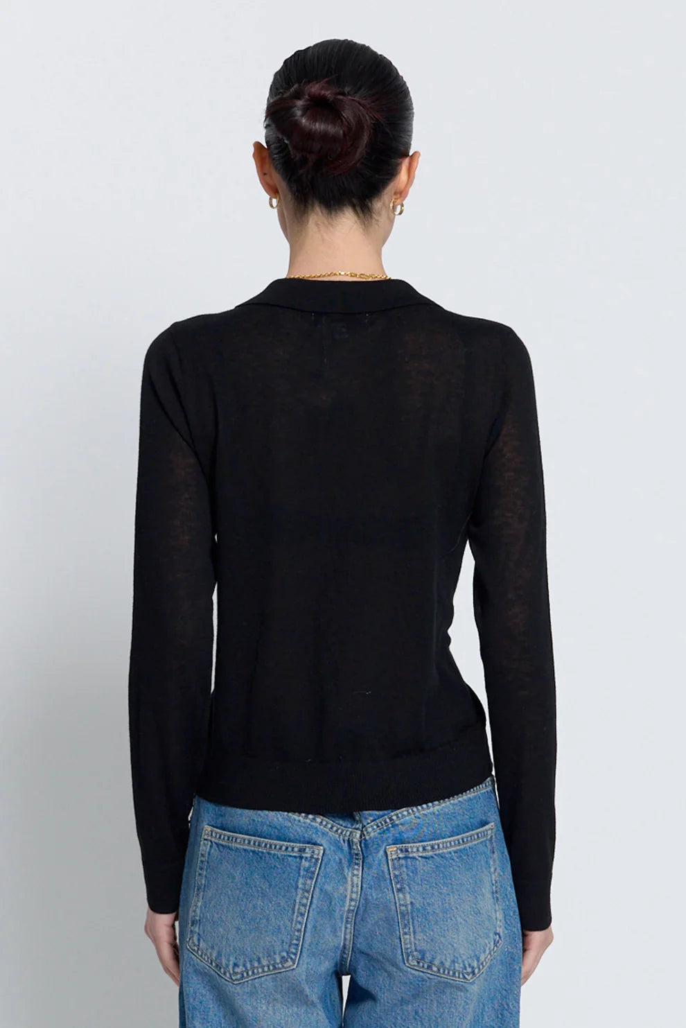 A woman viewed from the back wearing a black long-sleeve knit top and blue jeans against a light gray studio background.