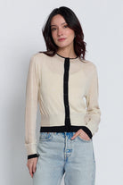 A woman wearing a light beige knit cardigan with black trim and a black button placket, paired with light-wash blue jeans, against a plain light gray studio background.