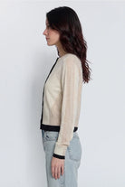 A woman shown in profile wearing a beige knit cardigan with black trim and light-wash jeans against a plain light gray studio background.