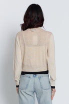A woman viewed from the back wearing a light beige knit cardigan with a black waistband and cuffs, paired with light blue jeans, against a plain light gray studio background.