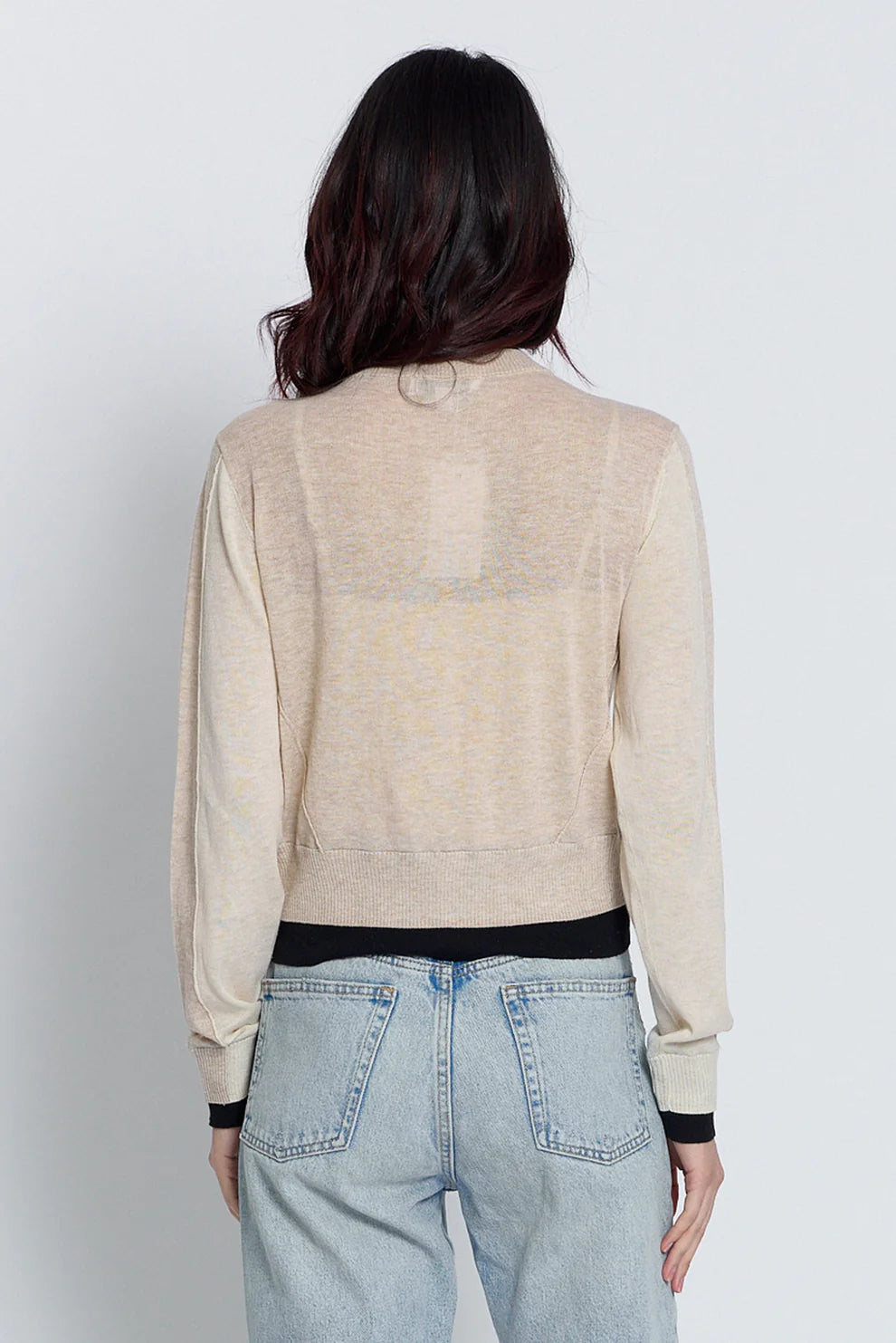 A woman viewed from the back wearing a light beige knit cardigan with a black waistband and cuffs, paired with light blue jeans, against a plain light gray studio background.