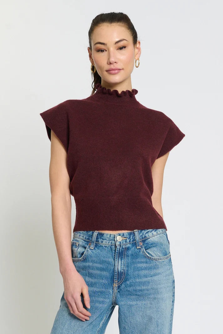 A woman modeling a sleeveless, high-neck ribbed knit top in deep burgundy with a ruffled trim at the neckline, paired with blue jeans, against a plain light gray background.