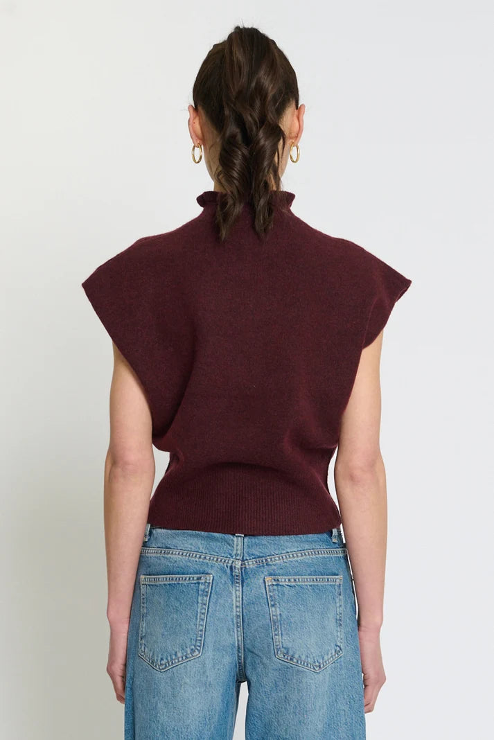 A woman viewed from the back wearing a sleeveless, mock-neck knit sweater in deep burgundy and light-wash blue jeans, set against a plain light gray studio background.