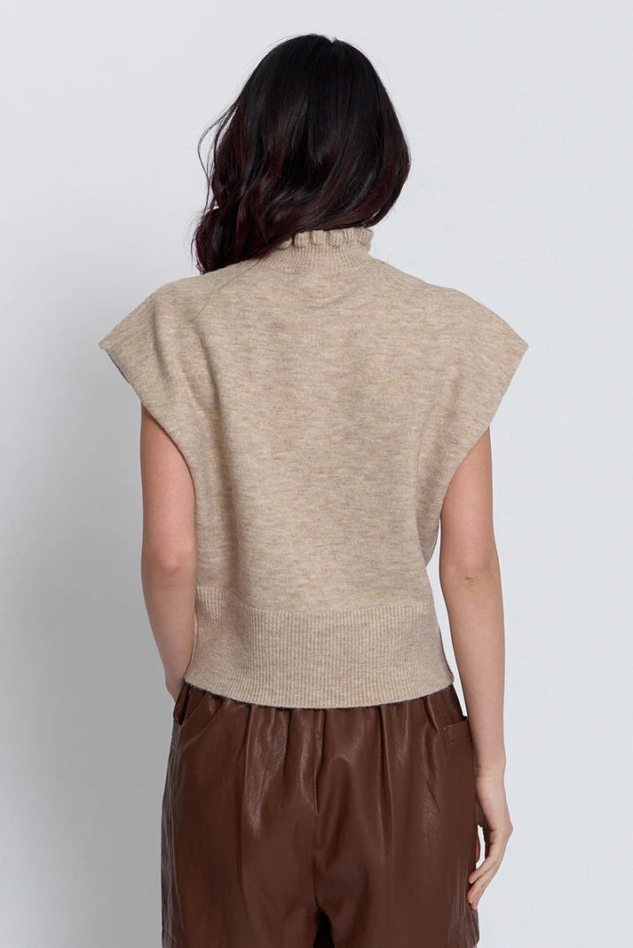 A woman shown from the back wearing a beige ribbed knit mock-neck sleeveless top and brown pants, photographed against a plain light gray studio background.