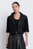 A woman modeling a black faux-leather cropped jacket vest over a black ribbed top and a black faux-leather pleated skirt, against a light gray studio background.