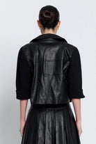 A model shown from the back wearing a black leather jacket with ribbed knit sleeves and a matching black pleated skirt, set against a plain light gray background.