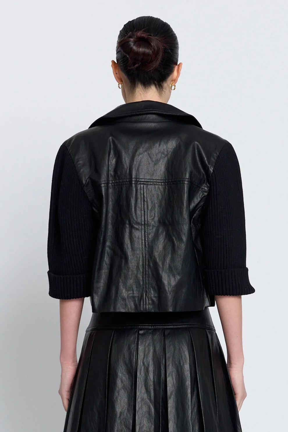 A model shown from the back wearing a black leather jacket with ribbed knit sleeves and a matching black pleated skirt, set against a plain light gray background.