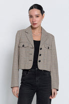 A woman modeling a cropped, long-sleeve plaid blazer in beige, brown, and black, worn over a black top and dark jeans against a light gray studio background.